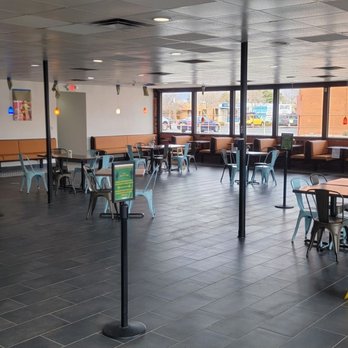 El Taco Tote - Dining area - Interior of El Taco Tote showing an open dining space with multiple tables and chairs, large windows along the exterior wall, and a counter area on the left. The floor is tiled, and there are some stanchions with signs. A yellow caution sign is placed on the floor, and a staff member is visible behind the counter. The space is well-lit with natural light from the windows. - Mexican Near Me - Albuquerque, New Mexico