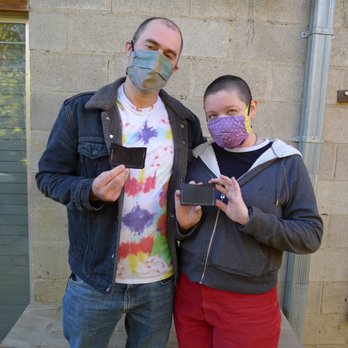 MAKE Santa Fe - Sweet couple took the sewing certification class. - A man and a woman stand outdoors in front of a beige brick wall. Both are wearing face masks; the man has a multicolored mask and the woman has a purple patterned mask. They each hold a small black card in their hands. The man is dressed in a black denim jacket with a fleece collar and a tie-dye T-shirt, while the woman wears a dark hoodie with a white lining and red pants. The setting appears to be outside a building with a window and metal piping visible. - Community Service/Non-Profit Near Me - Santa Fe, New Mexico