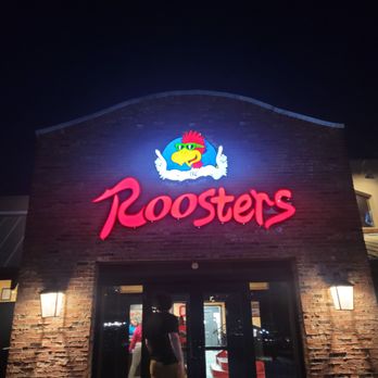 Roosters - The image shows the exterior of Roosters restaurant at night, with a brightly lit sign mounted on a brick building. The sign includes a cartoon rooster wearing sunglasses, with white wings and a red comb, above the word 'Roosters' in large, red neon script. The building has brick walls, and there are two wall-mounted lamps on either side of the entrance, which is visible at the bottom of the image. A person is seen standing near the entrance, partially silhouetted against the interior lighting. - Chicken Wings Near Me - Maumee, Ohio