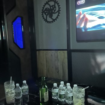 Stage Karaoke x Studios - The entrance to the studio room. - The image shows the interior of a karaoke room with a table holding several water bottles, a green bottle, and glasses with drinks. A crumpled tissue is on the table. A TV screen on the wall displays an image featuring a hand and water. The room has decorative gear-shaped wall art and a digital display with blue lighting. The setting suggests a casual entertainment environment. - Karaoke Near Me - Allston, Massachusetts
