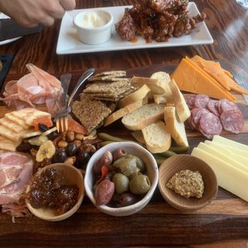Corner Bar and Kitchen - Antipasti platter - A cheese and charcuterie platter includes sliced cheeses, cured meats, assorted crackers, sliced bread, green and black olives, and small bowls of condiments and spreads. The platter is arranged on a wooden table, with additional dishes and drinks visible in the background, suggesting a shared dining experience. - Cocktail Bars Near Me - Rangeley, Maine