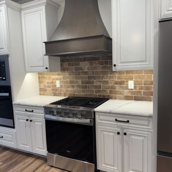 Denzal Custom Homes - The kitchen includes white raised-panel cabinets with black hardware, a stainless steel oven, and a brick backsplash behind the stove area. A large metallic range hood is mounted above the stove, and the countertop appears to be white with subtle veining. The flooring is wood or wood-like material, and there are electrical outlets on the backsplash wall. - General Contractors Near Me - Eynon, Pennsylvania
