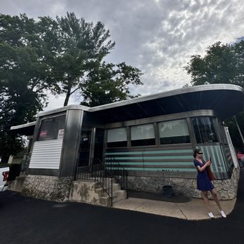 The Forked River Diner - classic diner architecture - A retro diner building with a curved roof, metal siding, and stone foundation, featuring neon signs and large windows. A woman stands outside, looking at her phone. - American Near Me - Forked River, New Jersey