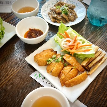 Pho Saigon - 5. Chao Tom - A rectangular white plate holds fried fish fillets garnished with chopped green onions, alongside fresh lettuce, shredded carrots, and pickled vegetables. Two small white bowls contain dipping sauces, one with a reddish chili sauce and the other with a light broth. Additional dishes include a plate of grilled meat and a glass of water on a dark wooden table, suggesting a Vietnamese restaurant setting. - Soup Near Me - Dover, Delaware