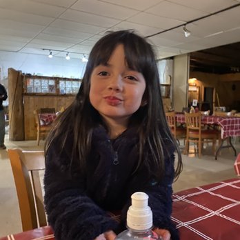 The Grill @ the MC - a little girl sitting at a table with a bottle of water - A girl with dark hair and a dark jacket is seated at a table in a restaurant, holding a bottle of hand sanitizer, with a background of wooden furniture and a rustic interior. - Bars Near Me - Questa, New Mexico