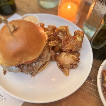 Sixty Vines - Burger and fingerling potatoes - A white plate holds a burger with a bun, melted cheese, and a beef patty, accompanied by fingerling potatoes. The table has a candle in a glass holder, water bottles, and glasses, indicating a dining setting at a restaurant. - New American Near Me - Miami, Florida