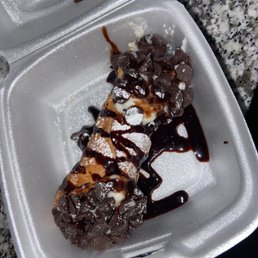 Cannoli - A cannoli pastry filled with cream, topped with chocolate chips, and drizzled with chocolate sauce, presented in a foam takeout container on a granite countertop. The dessert appears to be partially eaten or prepared for serving.