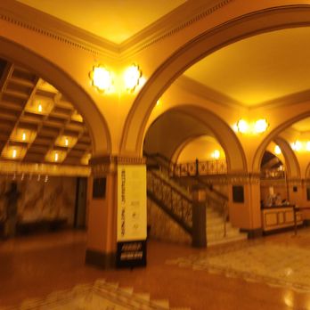 Auditorium Theatre - A photograph captures the interior of a historic theatre featuring ornate architectural details, including large arches and decorative moldings. The space is illuminated by warm yellow lighting from wall-mounted fixtures, creating a cozy atmosphere. A staircase with a decorative railing leads to an upper balcony, and the floor has a patterned design. The setting suggests a venue designed for performances or events, with a classic and elegant style. - Performing Arts Near Me - Chicago, Illinois