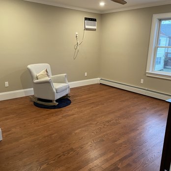 Murphy Construction - New bedroom pic 2 - The image shows a room with hardwood flooring and beige walls. There is a white armchair with a pillow placed on a small dark rug. A window on the right side provides natural light, and a wall-mounted air conditioner is visible on the wall. The ceiling has a modern wooden fan and recessed lighting. Baseboard heating runs along the bottom of the walls. - General Contractors Near Me - Oceanside, New York
