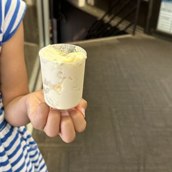 What Tea - Ice cream - A person is holding a small, clear cup of ice cream with a creamy texture, in front of a food court area. The background includes a black floor, some stairs, and a food stall with signage, indicating an indoor dining or shopping environment. - Bubble Tea Near Me - Boston, Massachusetts