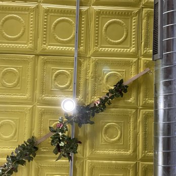 Raw Edge - Cheery ceiling! - The image shows a ceiling with yellow, ornate square panels, each decorated with detailed molding and circular motifs. A branch with dark green leaves and small flowers is attached to the ceiling, crossing the image diagonally. A bright light and a metallic duct are also visible, indicating an indoor setting with decorative and functional elements. - Flowers & Gifts Near Me - Chicago, Illinois