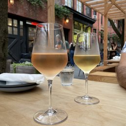 Rosé and white wine by the glass - Two wine glasses, one with rosé and one with white wine, are placed on a wooden table in an outdoor dining setting with a brick building and wooden pergola.