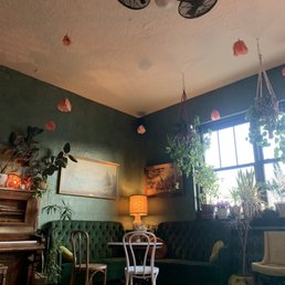 The image shows a cozy interior corner with vintage-style furniture, including a green tufted sofa, wooden chairs, and a side table with a lamp. Several potted plants hang from the ceiling and are placed near a large window, allowing natural light to fill the space. The walls are decorated with paintings, and the ceiling has hanging paper flowers and a ceiling fan with two small fans attached. The overall ambiance is warm and inviting, with a mix of natural and artificial lighting, and a variety of greenery adding to the decor.
