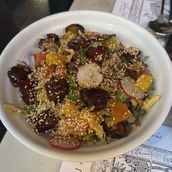 Eyval - Citrus Date & Cabbage Salad, lettuce, cabbage, orange, sesame seed, chili tahini dressing  18 - A mixed salad featuring radish slices, orange segments, dates, and sesame seeds, served in a white bowl on a table. - Persian/Iranian Near Me - Brooklyn, New York