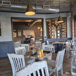 Dining area - A restaurant dining space featuring wooden tables, white chairs, pendant lighting, and a wine rack on the wall, with an open kitchen area in the background.