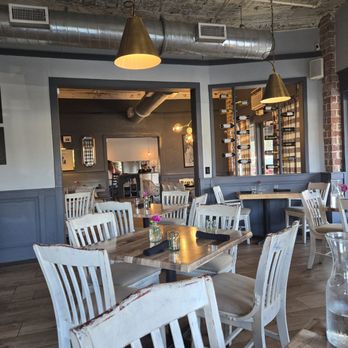 Crush XI - Dining area - A restaurant dining space featuring wooden tables, white chairs, pendant lighting, and a wine rack on the wall, with an open kitchen area in the background. - New American Near Me - Melbourne, Florida