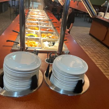 China Buffet - A buffet station featuring a variety of Asian dishes in metal trays, with stacked white plates and utensils in the foreground. The restaurant interior includes brick flooring, tables, and chairs, with warm lighting overhead. A staff member is visible in the background, and the buffet is arranged along a long counter with heat lamps overhead. - Chinese Near Me - Lima, Ohio