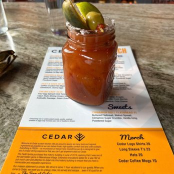 Cedar - A Bloody Mary cocktail in a glass jar with salt-rimmed edges, garnished with lime, green olives, and a pickle spear. The drink is on a restaurant menu at Cedar in Covington, KY, with a glass of water and sunglasses nearby. The menu features various food and drink options, and the background shows a bar area. - Breakfast & Brunch Near Me - Covington, Kentucky