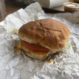 LTP burger with cheese with grilled onions - A burger with a toasted bun, melted cheese, a slice of tomato, and lettuce is placed on crumpled white paper. The background shows a fast-food environment with a paper cup, a small container of sauce, and other food items on a table. The burger appears to be a typical American-style fast-food sandwich.