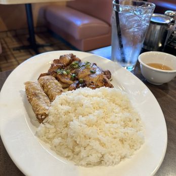 Việt kitchen - 56. Rice with Grilled Chicken & Egg Roll Plate - A white oval plate holds a serving of steamed white rice, grilled chicken with green onions, and two egg rolls. The plate is on a dark wooden table in a restaurant setting. Next to the plate, there is a glass of water with ice and a straw, a small bowl of tea, and a metal teapot. The background shows booth seating with brown cushions and wooden tables. - Vietnamese Near Me - Albuquerque, New Mexico