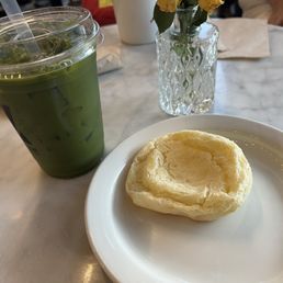 Matcha lemonade and a tasty  sticky cheese treat ( I forgot the name) but they bake it in store. - A plastic cup with a clear lid and straw contains a green matcha lemonade with ice cubes. Next to it, a white plate holds a baked cheese pastry with a golden, flaky crust. A small, textured glass vase with yellow flowers is also on the marble table, with a napkin and part of a person's arm visible in the background.