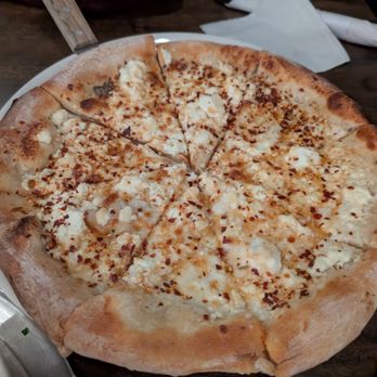 ZA - Goat pizza - A cheese pizza with a thick, golden-brown crust and melted cheese topping, cut into slices, is placed on a dark wooden table. The pizza appears to have some chili flakes or red pepper seasoning on top. A white napkin and a utensil are visible in the background, along with a person wearing a dark-colored shirt. - Pizza Near Me - Elm Grove, Wisconsin