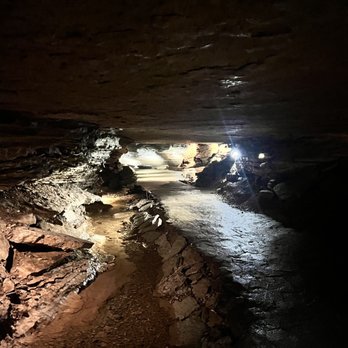 War Eagle Cavern - A dark, underground cavern with a rocky pathway and a small stream running through it, illuminated by a distant light source. - Historical Tours Near Me - Rogers, Arkansas