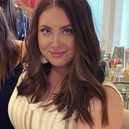 Went short with highlights - A woman with long, wavy brown hair and blue eyes is smiling at the camera in an indoor setting with other people and colorful items in the background.