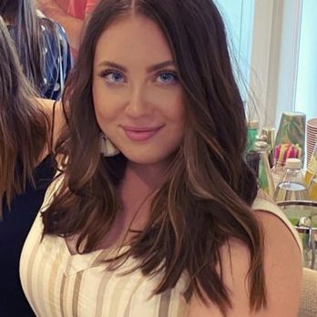 Salon Hype - Went short with highlights - A woman with long, wavy brown hair and blue eyes is smiling at the camera in an indoor setting with other people and colorful items in the background. - Hair Salons Near Me - La Grange, Illinois