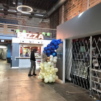 Sherman Phoenix - Inside there are a number of different food and beverage options. - Colorful balloon arrangements are positioned outside a pizza stand within an indoor marketplace, featuring exposed brick walls and industrial ceiling elements. - Food Near Me - Milwaukee, Wisconsin