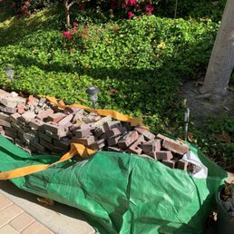A pile of bricks and construction debris is placed on a green tarp along a sidewalk. The debris includes various bricks and broken materials. Behind the debris, there is a garden area with green bushes, flowering plants, and a tree with a visible trunk. Small landscape lights are installed along the garden edge, and fallen leaves are scattered on the ground.