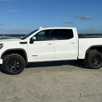 Infinity Conversions - A white pickup truck is positioned on a sandy beach, facing left, with the ocean waves and a partly cloudy sky in the background. The truck has black wheels and tinted windows, and the scene suggests a coastal or recreational setting. - Car Stereo Installation Near Me - Round Rock, Texas