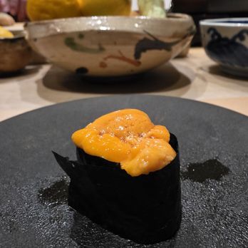 Hiden - Uni hokkaido - A piece of uni (sea urchin) sushi, wrapped in nori (seaweed), is placed on a black plate. The uni has an orange color and a creamy texture. In the background, there are bowls with decorative patterns and a bowl containing lemons and other fruits, suggesting a dining environment with Japanese cuisine elements. - Japanese Near Me - Miami, Florida