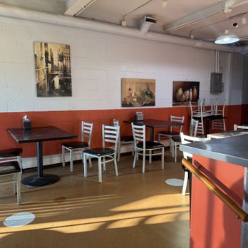 Dylan’s Pizza - Dining in? - A restaurant interior features several tables and chairs against a white and red wall, with artwork hanging above. Sunlight casts shadows on the floor. - Pizza Near Me - Hull, Massachusetts