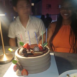 A chocolate birthday cake decorated with strawberries, mint leaves, and chocolate curls is placed on a white board on a table. Four lit candles are on top of the cake. Two people, a man and a woman, are sitting behind the cake, smiling and celebrating. The setting appears to be outdoors at night, with other people and buildings visible in the background.