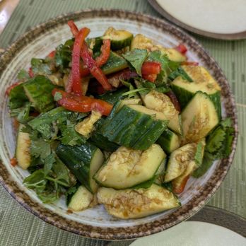 Laghman Express - A ceramic bowl contains a vegetable salad made of chopped cucumbers, red bell peppers, and fresh herbs such as cilantro. The salad appears to be dressed with a light sauce or dressing. Two additional bowls of sauce or dressing are partially visible on the table, which has a green woven placemat. The setting suggests a casual dining environment, possibly at a restaurant specializing in fresh, healthy dishes. - Halal Near Me - Brooklyn, New York
