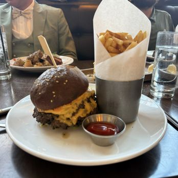 Agustin Kitchen - Crust Burger - A burger with a dark bun, cheese, and toppings is served with a side of fries in a paper cone and ketchup, on a white plate. - New American Near Me - Tucson, Arizona