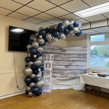 Balloonantics - Arch for group pics - A balloon arch made of navy, silver, and metallic balloons is displayed in a room with a wooden floor and a backdrop. The room has a window, a TV, and a table with food. - Balloon Services Near Me - Saugus, Massachusetts
