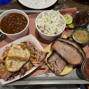 2 Jerks BBQ - 2 Meat Platter with brisket and pulled pork, beans and cole slaw sides - A tray displays a barbecue meal with sliced brisket, pulled pork with gravy, baked beans, coleslaw, and pickles. There are small containers of barbecue sauce and mustard, and a spoon is placed on the tray. The meal is served on a gray tray with a paper liner, set on a wooden table. - Barbeque Near Me - Raynham, Massachusetts