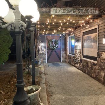 Blackhawk Steak Pit - Beautiful, ambience and entrance. - Nighttime scene of the entrance to Blackhawk Steak Pit, with a sign above the doorway. The door is decorated with a wreath, and string lights are hung overhead, creating a festive atmosphere. A vintage-style lamp post with multiple globes illuminates the walkway, which is lined with potted plants. The building has stone and siding exterior walls, and a window with a landscape photograph. The setting suggests a cozy, inviting ambiance for diners. - Steakhouses Near Me - Oregon, Illinois