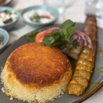 Joon - A ceramic plate holds a Persian-style meal including a large, golden-brown rice cake, a grilled meat skewer, and fresh herbs with sliced tomatoes and onions. The background shows small bowls with side dishes and a glass of water, indicating a dining setting at Joon in Vienna, VA. - Persian/Iranian Near Me - Vienna, Virginia