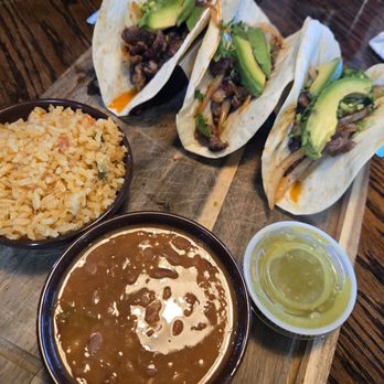 Daddy Shark - Rib-eye Tacos - Three beef tacos with sliced avocado and cilantro are placed on a wooden serving board. The tacos are made with soft tortillas and filled with cooked beef. A bowl of rice and a bowl of beans are positioned in front of the tacos, along with a small cup of green sauce. The setting appears to be a restaurant with a dark wooden table surface. - Seafood Near Me - Clinton, Mississippi