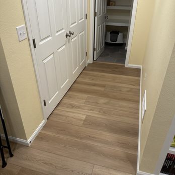 Simply Floors & Painting - New Lyrus LVP Flooring - Mesa is the color. - The image shows a hallway with light wood flooring, white double doors, and an open closet with shelves and a small appliance. - Flooring Near Me - Centennial, Colorado