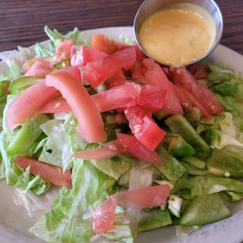 Nick's Cafe - Dinner side salad - A plate of fresh side salad includes chopped lettuce, sliced tomatoes, and cucumber pieces. A small metal cup of salad dressing is placed on the plate. The salad is served on a white dish, with a wooden table surface visible in the background. - Breakfast & Brunch Near Me - Toledo, Ohio