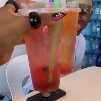 Soule' Coffee + Bubble tea - Build your own boba teas (red one): Lemonade base, mixed fruit, dragonfruit syrup, strawberry boba - A person is holding two large bubble tea drinks with colorful straws, positioned in front of a man wearing a cap with a tropical design and the word 'Details.' The drinks are in clear plastic cups, showing the beverage with fruit and jelly toppings. The setting appears to be inside a beverage shop, with bottled water stacked in the background. The person holding the drinks has painted nails and is wearing rings and a black bracelet. - Coffee & Tea Near Me - Jackson, Mississippi