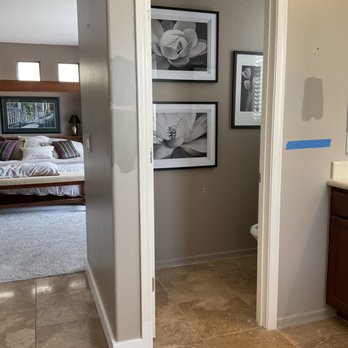 Tru Builders - BEFORE Toilet Room - The image shows a doorway with white trim leading into a small room. Inside, three black-and-white floral photographs are hung on the wall, with two larger pictures positioned horizontally and one smaller picture vertically. The room has beige walls and a tiled floor. To the left, part of a bedroom with a bed, pillows, and a framed picture above the headboard is visible. - General Contractors Near Me - Surprise, Arizona