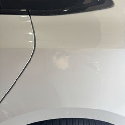 Unable to find correct paint for touch up which looked off - The image depicts a white car with a noticeable dent and scratch on the side panel near the door. The car has a black window at the top and a tire at the bottom, with reflections on the car's surface indicating indoor lighting. The damage appears to be superficial, affecting the paint and surface of the vehicle.