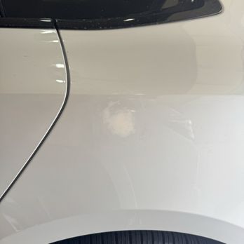 Bumper Buddies - Unable to find correct paint for touch up which looked off - The image depicts a white car with a noticeable dent and scratch on the side panel near the door. The car has a black window at the top and a tire at the bottom, with reflections on the car's surface indicating indoor lighting. The damage appears to be superficial, affecting the paint and surface of the vehicle. - Mobile Dent Repair Near Me - Phoenix, Arizona