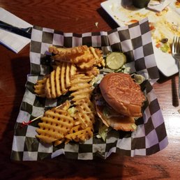 Damn Burger and fries - A burger with a toasted bun, lettuce, and other toppings is placed on checkered paper on a wooden table. Accompanying the burger are waffle-cut fries and sliced pickles. In the background, there are additional plates with food, including a plate with guacamole and remnants of a meal, and utensils are visible on the table.