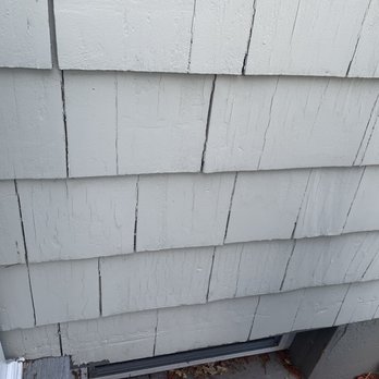 Excel Painting - The image depicts a section of an exterior wall with horizontal siding made of wood or fiber cement, painted in a light color. Visible cracks and weathering are present on the siding, indicating age or exposure to the elements. The bottom part of the wall shows a small section of a window or door frame and some ground debris. The focus is on the wall's surface and condition. - Painters Near Me - Attleboro, Massachusetts